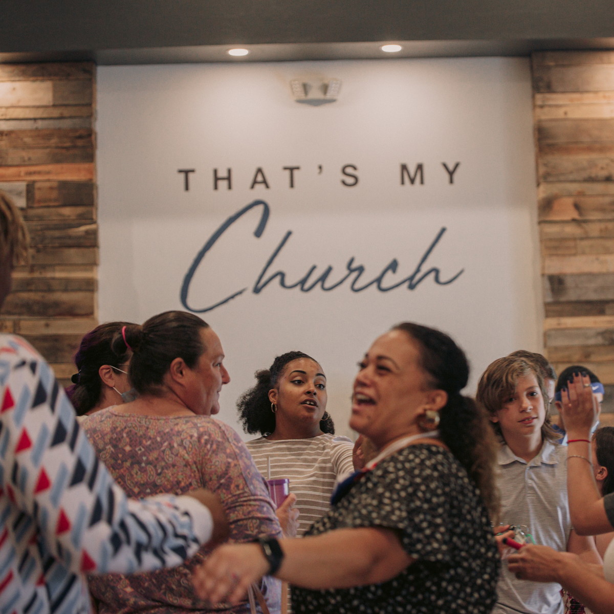 A group of people socialize in a warmly lit room with a Thats My Church sign on the wall.